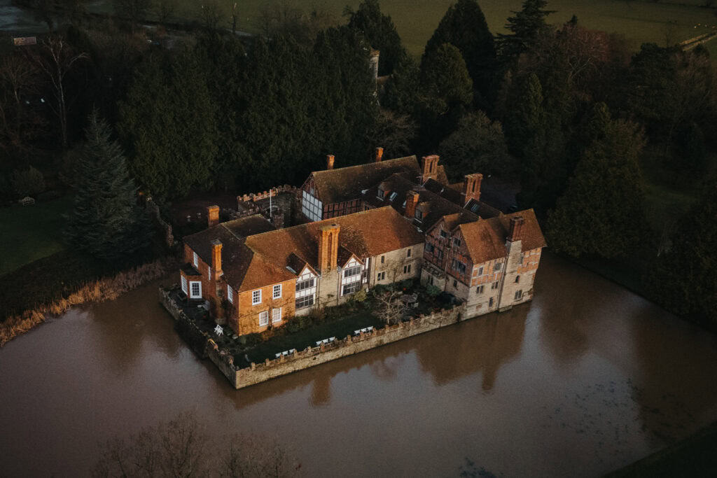 A stunning aerial view of Birtsmorton Court, a historic moated manor house nestled in the Cotswolds countryside. The medieval red-brick and timber-framed architecture, surrounded by a tranquil moat, is beautifully illuminated by the soft winter light. The lush greenery and towering trees provide a picturesque backdrop, making it an enchanting wedding venue, especially in the colder months.