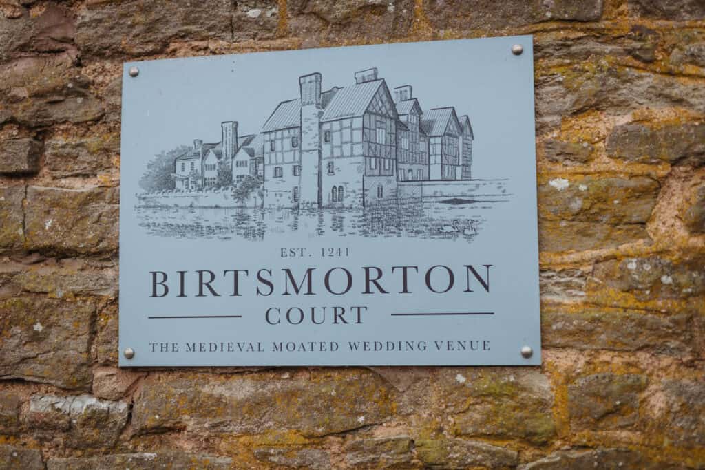 A close-up of a sign mounted on a rustic stone wall, welcoming visitors to Birtsmorton Court. The sign features an elegant black-and-white illustration of the historic moated manor house, along with the text ‘Birtsmorton Court, Est. 1241 – The Medieval Moated Wedding Venue.’ The textured stone backdrop adds to the timeless charm of this renowned Cotswolds wedding location.