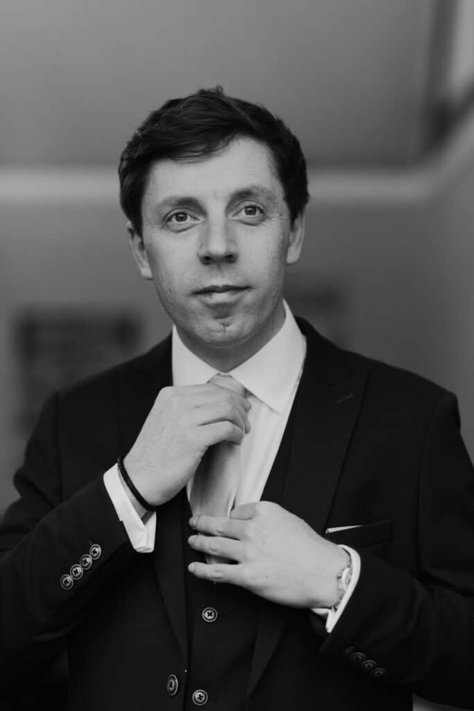 A black and white portrait of Lewis, the groom, adjusting his tie as he prepares for his wedding day at Birtsmorton Court. Captured in a candid moment, his focused expression reflects anticipation and emotion. Dressed in a sharp three-piece suit, the fine details of his attire, including the cuff buttons and wrist accessories, add to the elegance of this timeless wedding preparation shot.