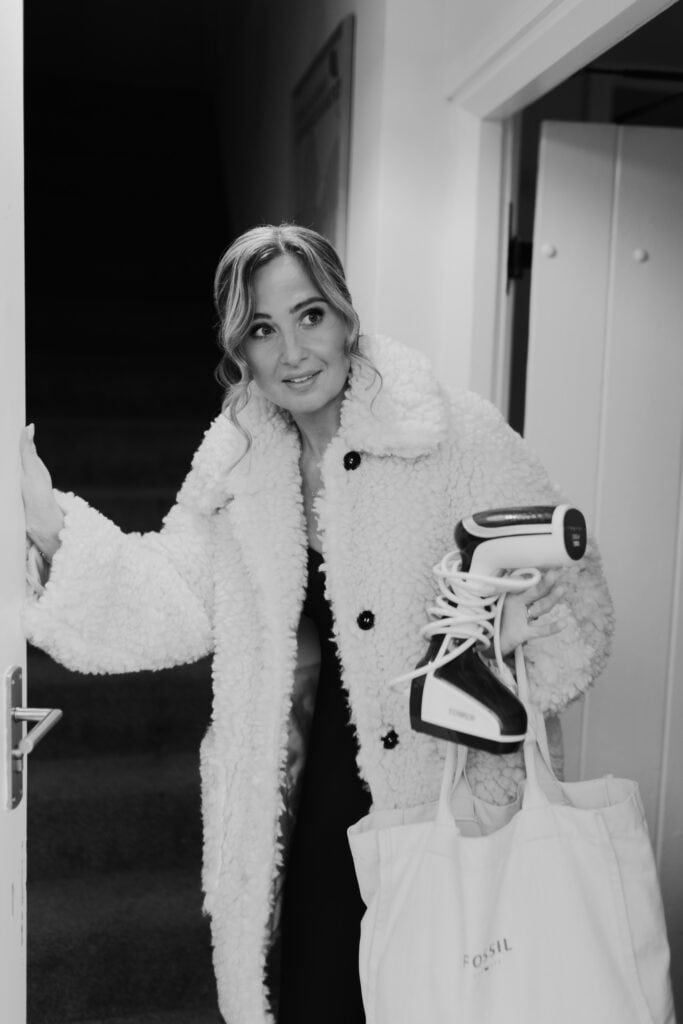 A black and white candid portrait of Jess, the bridesmaid, arriving during wedding preparations at Birtsmorton Court. Wrapped in a cosy faux-shearling coat, she holds a garment steamer and a Fossil tote bag, exuding a mix of excitement and focus.