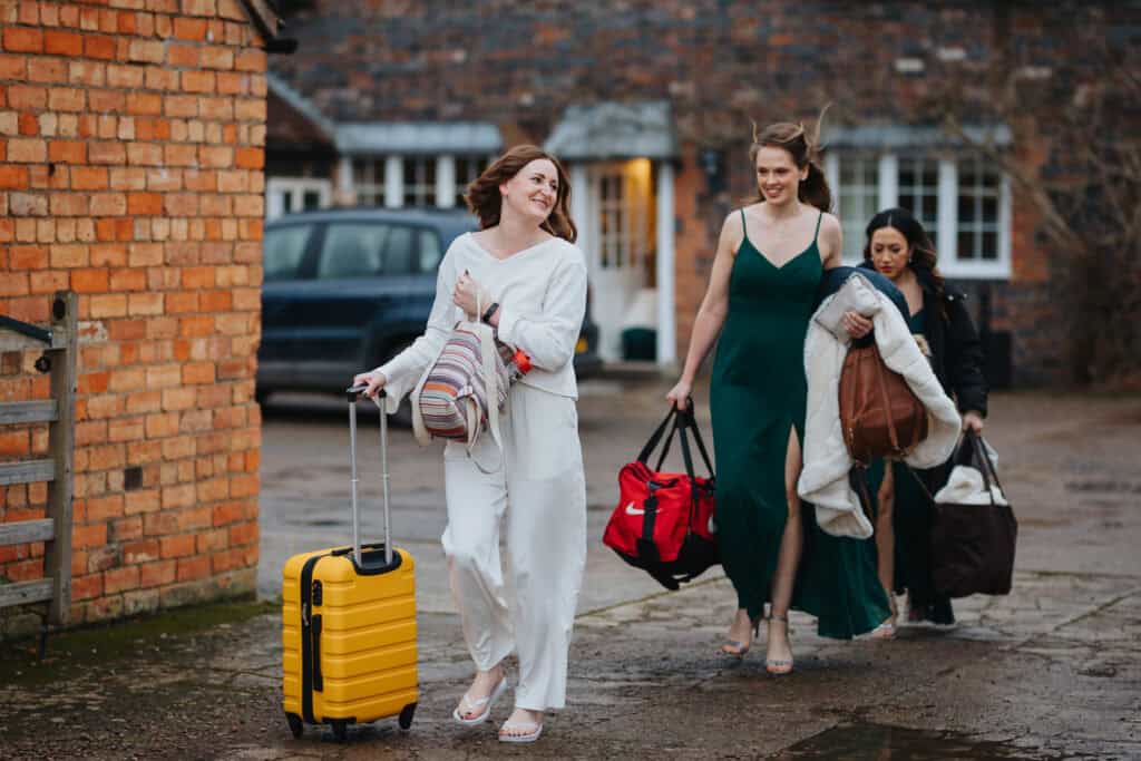 Bride Katie arrives at Birtsmorton Court with her bridesmaids, carrying bags and a bright yellow suitcase, ready for the wedding preparations. Dressed in a relaxed white loungewear set, she smiles joyfully as her bridesmaids, elegantly dressed in deep green gowns, follow behind with coats and overnight bags. The candid moment captures the excitement and anticipation of the day in the charming rustic courtyard setting.