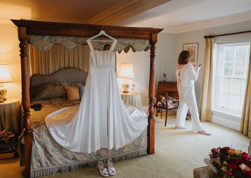 A beautifully composed bridal preparation scene at Birtsmorton Court. Katie, the bride, stands near a window in soft white loungewear, absorbed in her phone, while her elegant wedding dress hangs from the four-poster bed, its flowing fabric draped across the luxurious bedding. A pair of white heels sits neatly below, completing the bridal look. Warm lighting and classic décor create an intimate and serene atmosphere, capturing the anticipation of the wedding day.