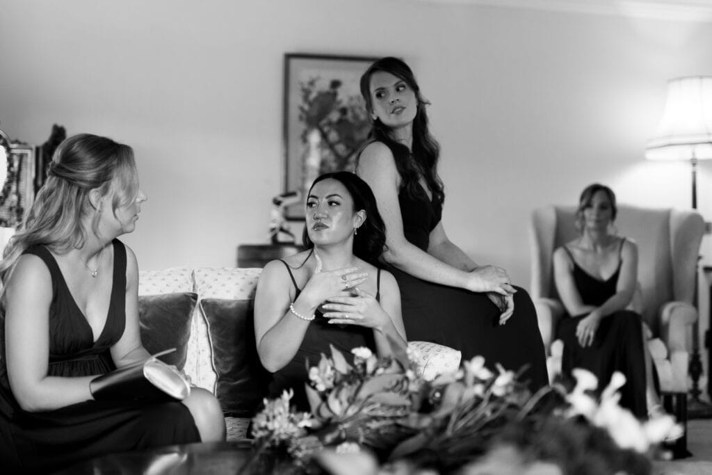 A timeless black and white moment of the bridesmaids gathered in a cosy lounge at Birtsmorton Court. Dressed in elegant dark gowns, they engage in conversation, each displaying a mix of anticipation and emotion. One bridesmaid adjusts her jewellery while another listens attentively, as soft lighting and classic furnishings set the perfect atmosphere for a heartfelt pre-wedding moment.