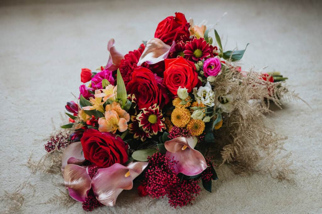 A vibrant and elegantly arranged bridal bouquet featuring rich red roses, deep burgundy calla lilies, golden chrysanthemums, and delicate pink and white blooms. The lush arrangement is accented with wispy gold foliage, creating a luxurious and warm colour palette perfect for a winter wedding at Birtsmorton Court. The bouquet rests on a soft, neutral background, highlighting its intricate textures and seasonal charm.