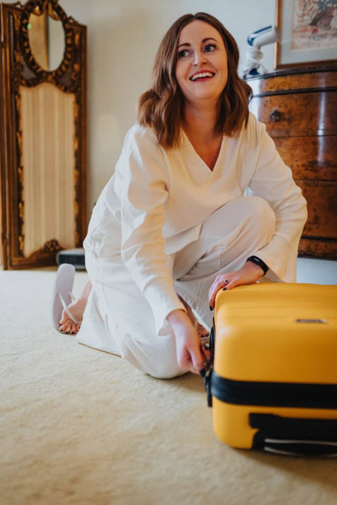Bride Katie, dressed in relaxed white loungewear, kneels on the floor while unzipping her bright yellow suitcase during wedding preparations at Birtsmorton Court. She smiles joyfully, capturing the excitement of the morning. The elegant vintage mirror and wooden dresser in the background add a classic touch to this candid and heartwarming moment.