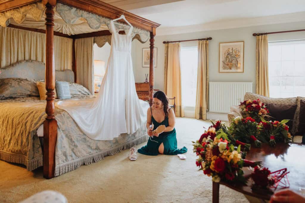 A bridesmaid, dressed in an elegant deep green gown, kneels on the floor as she prepares for the wedding day at Birtsmorton Court. She carefully adjusts her shoes beside a beautifully draped wedding dress hanging from a four-poster bed. The soft lighting, classic furnishings, and fresh bouquets of red and white flowers on the table create a warm and intimate pre-wedding scene.