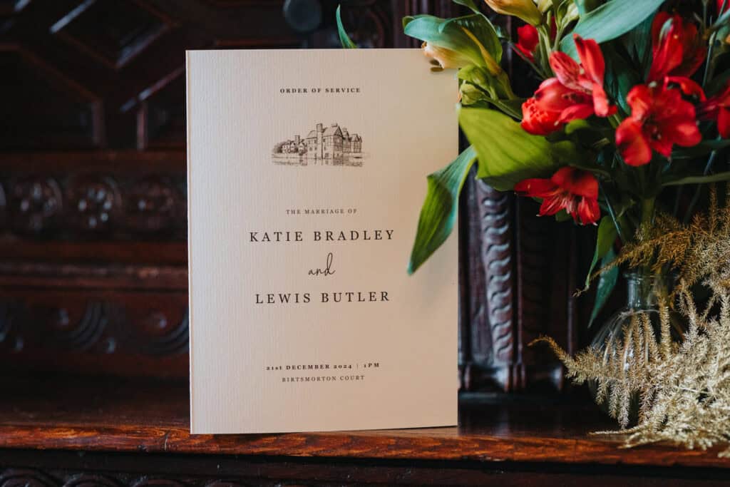 A beautifully designed wedding order of service card for the marriage of Katie Bradley and Lewis Butler, set against an ornate wooden backdrop at Birtsmorton Court. The elegant card features a delicate sketch of the historic venue and refined typography, announcing the wedding date as 21st December 2024 at 1 PM. Beside it, a floral arrangement of red and gold blooms adds a warm, festive touch to the intimate winter wedding setting.