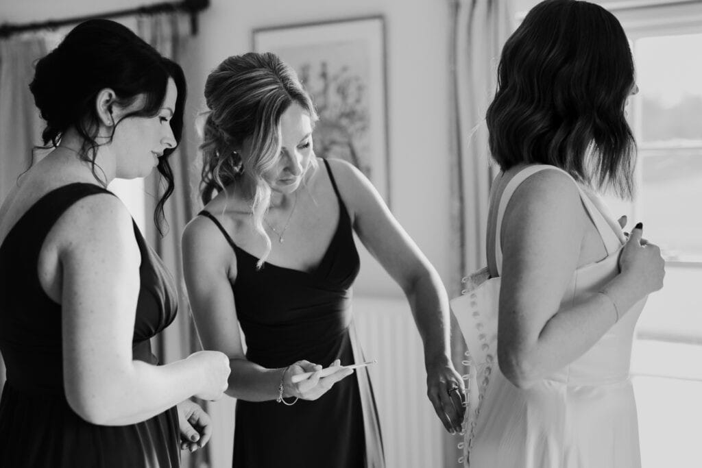 A black and white photograph capturing an intimate wedding morning moment at Birtsmorton Court. The bride, dressed in a sleek satin gown with delicate button detailing, stands near a sunlit window as two bridesmaids, wearing elegant dark dresses, help fasten her dress. One bridesmaid carefully secures the buttons, while the other observes with a warm expression. The soft natural light and timeless monochrome tones highlight the anticipation and closeness of the moment.