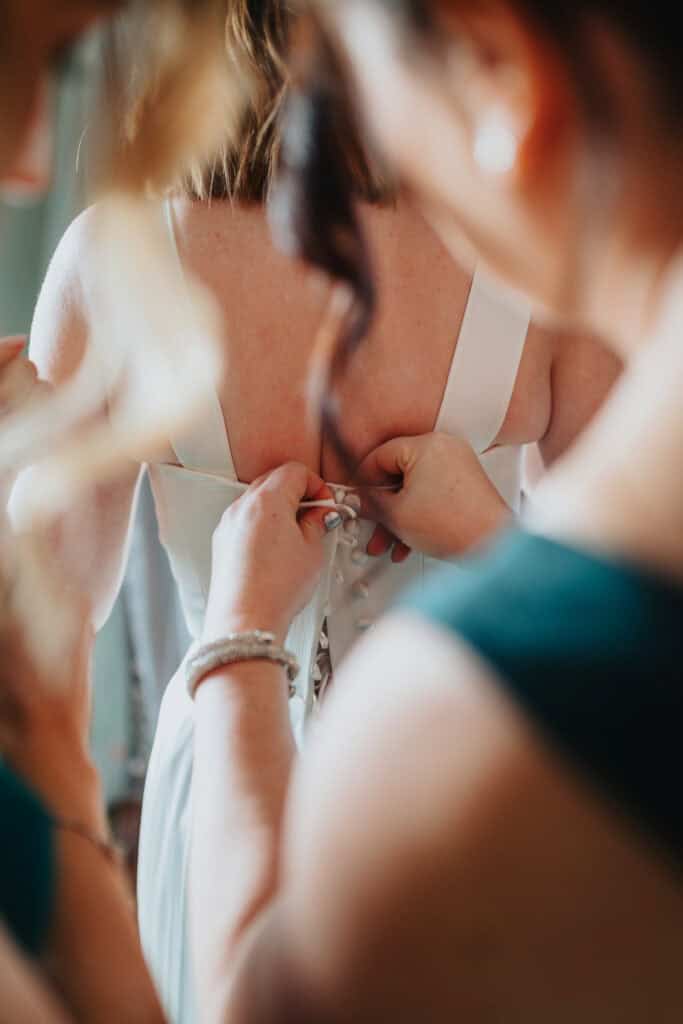 A close-up, intimate moment as the bride's wedding dress is carefully fastened by her bridesmaids at Birtsmorton Court. Their hands delicately secure the elegant row of buttons along the back of the gown, highlighting the intricate details of the dress. The soft lighting and blurred foreground create a warm, emotional atmosphere, capturing the excitement and significance of the wedding preparations.