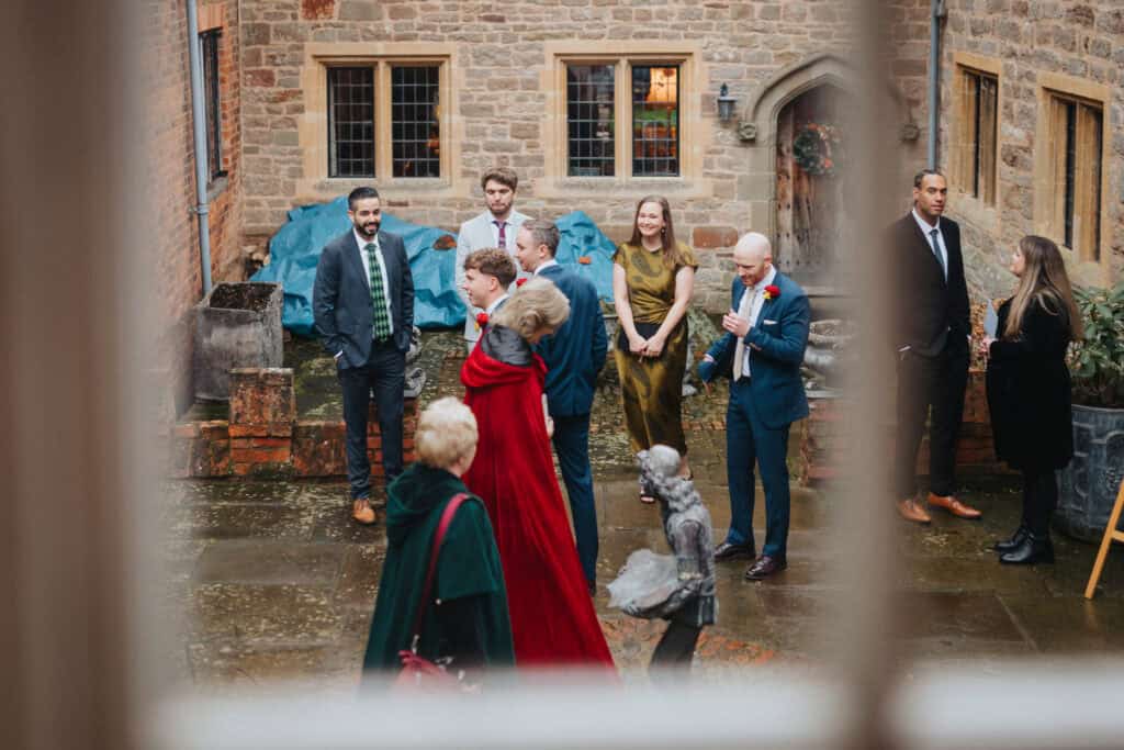 Wedding guests gather in the historic courtyard of Birtsmorton Court during a winter wedding celebration. Captured through a window, this candid moment showcases elegantly dressed guests in suits and formal attire, engaging in conversation before the ceremony. The rustic stone walls and festive greenery enhance the charm of this Cotswolds wedding venue, making it an idyllic setting for natural and documentary-style wedding photography.