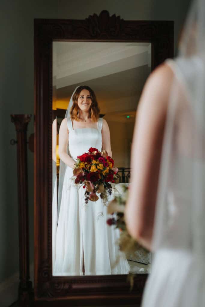Bride Katie admires her reflection in an ornate mirror at Birtsmorton Court on her wedding day. Dressed in an elegant white gown with a flowing veil, she holds a stunning bouquet of red and gold flowers, perfectly complementing her winter wedding theme. This bridal portrait captures a timeless moment of joy and anticipation, framed by soft natural light and luxurious surroundings. A perfect example of elegant documentary wedding photography in the Cotswolds.