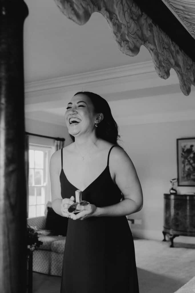 Bridesmaid laughing joyfully while holding a perfume bottle during wedding preparations at Birtsmorton Court. Captured in elegant black and white, this candid bridal party moment showcases natural emotion and excitement before the ceremony. Featuring soft lighting and a luxurious four-poster bed, this documentary-style wedding photography highlights the beauty of authentic wedding day emotions in a historic Cotswolds venue.