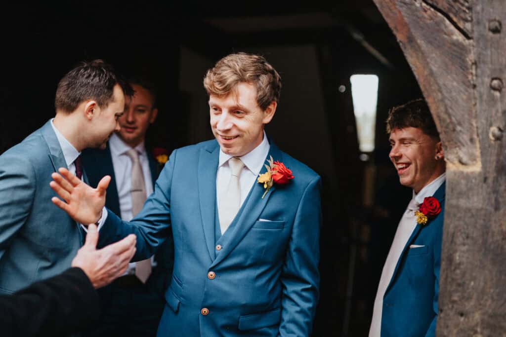 Groom and groomsmen share a joyful moment before the ceremony at Birtsmorton Court. Dressed in stylish blue suits with red and gold boutonnieres, they exchange smiles and greetings, capturing the excitement of the wedding day. This natural and candid wedding photography highlights the groom’s anticipation and the camaraderie of the bridal party, set against the rustic charm of this historic Cotswolds wedding venue.