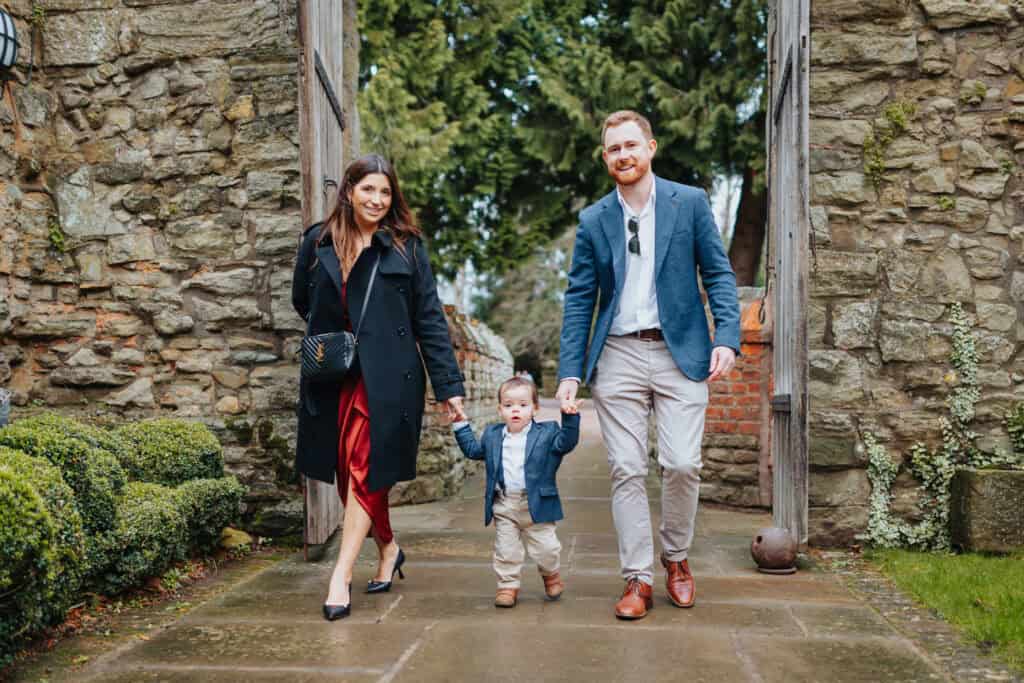 Stylish wedding guests arrive at Birtsmorton Court, walking hand-in-hand with their young child through the historic stone gate. The elegantly dressed couple, with the mother in a red satin dress and black coat and the father in a navy blazer, radiate joy as their little one, dressed smartly in a mini suit, takes confident steps.