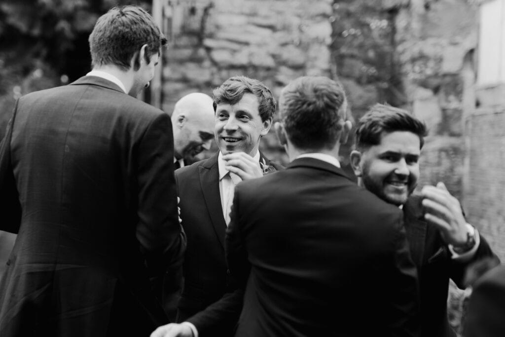 Candid black and white wedding photography of the groom sharing a joyful moment with his groomsmen at Birtsmorton Court. Surrounded by friends, he smiles as final preparations take place before the ceremony. This documentary-style wedding photo captures natural laughter, friendship, and anticipation at a stunning Cotswolds wedding venue, ideal for couples seeking relaxed and timeless wedding photography.