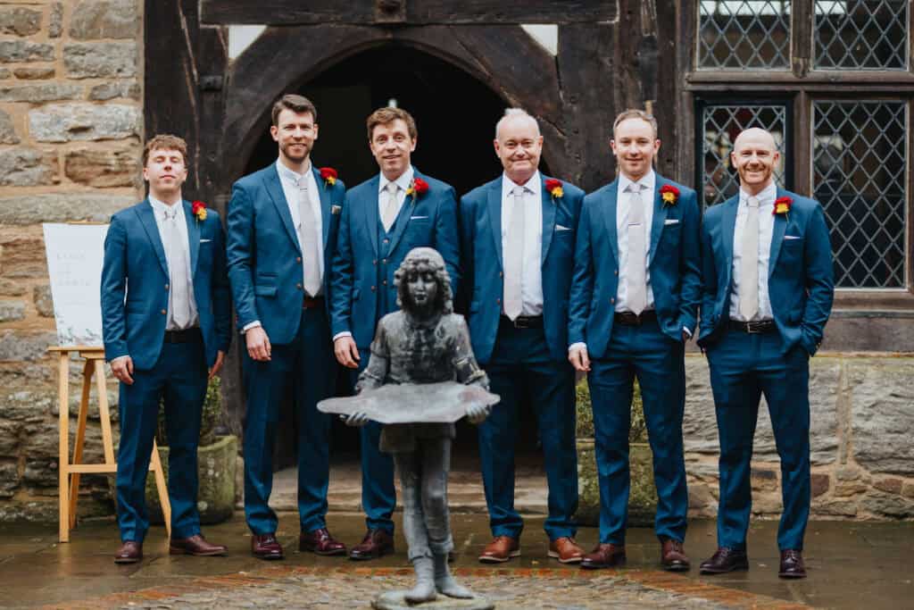 Groom and groomsmen pose in matching blue suits with red and gold boutonnieres at Birtsmorton Court, a historic Cotswolds wedding venue. Standing in front of the Tudor-style entrance, this classic wedding portrait captures the groom and his closest friends and family before the ceremony. This stylish and relaxed groomsmen group shot is perfect for couples seeking elegant and natural wedding photography with a timeless feel.