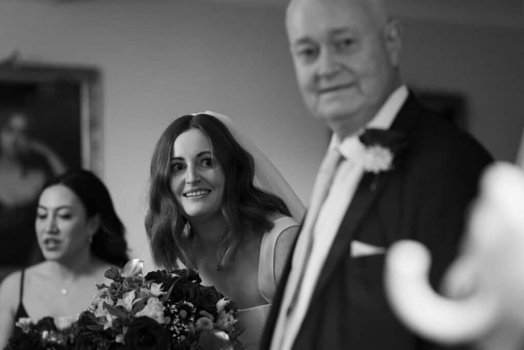 Bride Katie, holding her bouquet, shares a heartfelt moment with her father before walking down the aisle at Birtsmorton Court. Captured in elegant black and white, this emotional wedding morning portrait highlights the love and anticipation before the ceremony. A bridesmaid stands in the background, adding to the intimate and timeless atmosphere