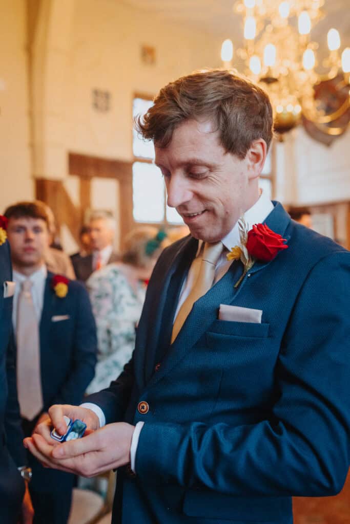 Groom Lewis at Birtsmorton Court, holding the wedding rings with a smile just moments before the ceremony. Dressed in a tailored navy-blue suit with a red rose boutonnière, he stands in the historic wedding venue’s candlelit setting, surrounded by groomsmen and guests. A heartfelt and intimate moment captured during this winter wedding in the Cotswolds.