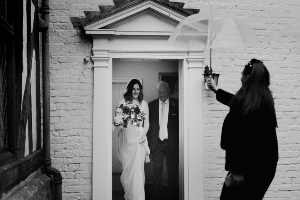 Bride Katie steps out with her father on a winter wedding day at Birtsmorton Court, holding a stunning bouquet of red and white flowers. A bridesmaid shelters them with a clear umbrella, adding a touch of romance to this timeless black and white wedding photography moment. Capturing the elegance and emotion of a Cotswolds wedding.