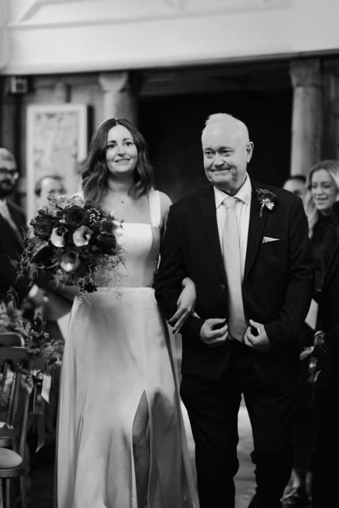 Bride Katie walks down the aisle with her father at Birtsmorton Court, a stunning winter wedding venue in the Cotswolds. Captured in timeless black and white, this emotional moment showcases her elegant bridal gown, a lush bouquet of red and blush flowers, and the proud father of the bride. A beautiful documentary-style wedding photograph preserving a cherished memory.