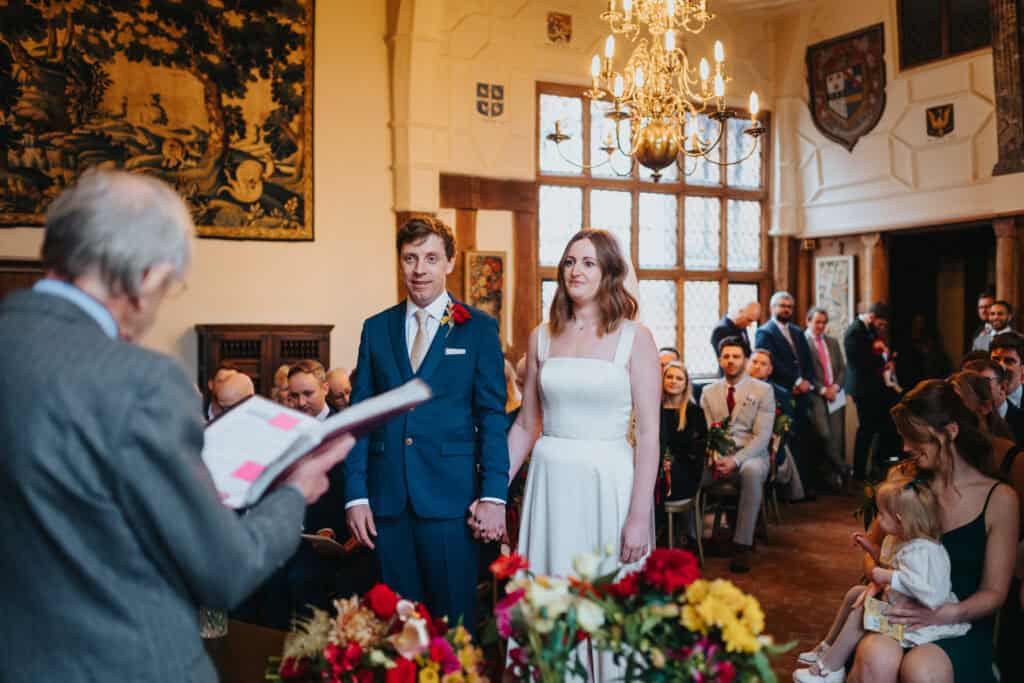 Bride and groom standing hand-in-hand during their wedding ceremony at Birtsmorton Court, a stunning Cotswolds winter wedding venue. The historic setting, warm candlelit ambiance, and floral decorations create a romantic atmosphere as the couple exchange vows in front of family and friends. Captured by a professional wedding photographer in Buckinghamshire, specialising in timeless and emotional wedding moments.
