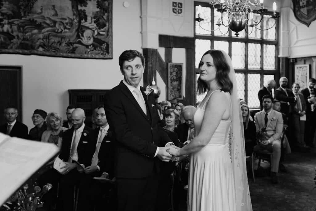 Black and white wedding ceremony moment at Birtsmorton Court, with the bride and groom exchanging vows in a historic, timber-framed setting. The groom holds the bride’s hands as they stand before family and friends, with natural light streaming through the grand windows. A timeless and emotional moment captured by a Buckinghamshire wedding photographer, specialising in storytelling wedding photography in the Cotswolds and beyond.