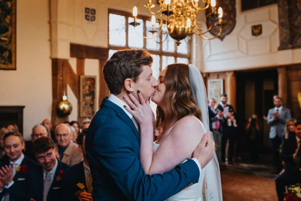 Bride and groom share their first kiss as a married couple at Birtsmorton Court, a stunning historic wedding venue in the Cotswolds. Captured in a beautifully lit ceremony room with timber beams and grand chandeliers, the newlyweds embrace as guests applaud in the background. A perfect moment from a romantic winter wedding, photographed by a Buckinghamshire wedding photographer specialising in authentic and emotive wedding photography.