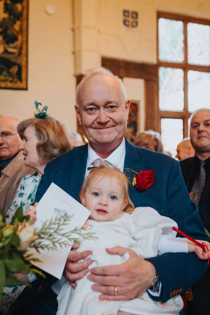 Heartwarming moment the father of the bride holding a young child during a beautiful winter wedding at Birtsmorton Court. Captured in the historic ceremony room, this touching family scene showcases the joy and love shared on the special day. The grandfather, dressed in a navy blue suit with a red rose boutonniere, holds the wedding order of service while smiling proudly.