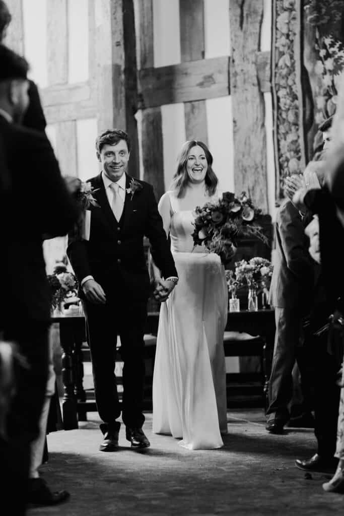 Just married! A joyful black and white wedding photograph of the bride and groom walking hand in hand down the aisle after saying 'I do' at Birtsmorton Court, a stunning winter wedding venue in the Cotswolds. The bride beams with happiness, holding a romantic bouquet of deep red and blush florals, while the groom looks overjoyed in a classic navy suit. Capturing raw emotions and timeless moments, this wedding photography perfectly encapsulates the love and excitement of the day.