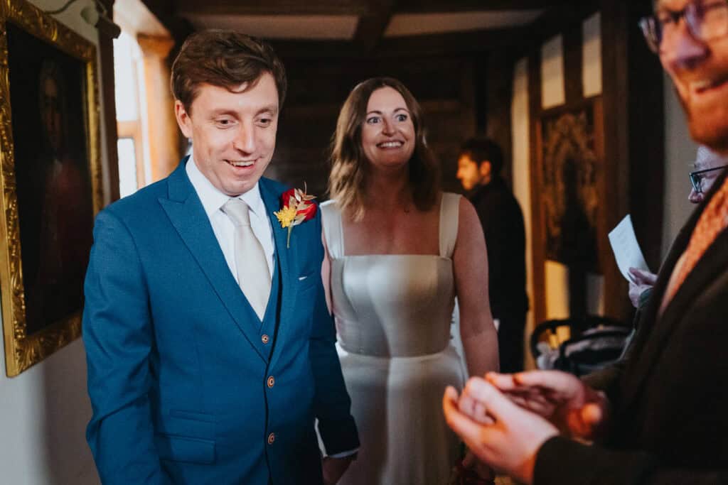 Just married! A candid wedding photography moment at Birtsmorton Court, capturing the bride and groom’s joyful expressions as they greet guests after their ceremony. The groom looks dapper in a tailored blue suit with a red and gold boutonnière, while the bride stuns in a timeless square-neck wedding dress. Set in a historic Cotswolds wedding venue, this heartwarming image highlights the excitement and love of their winter wedding celebration.