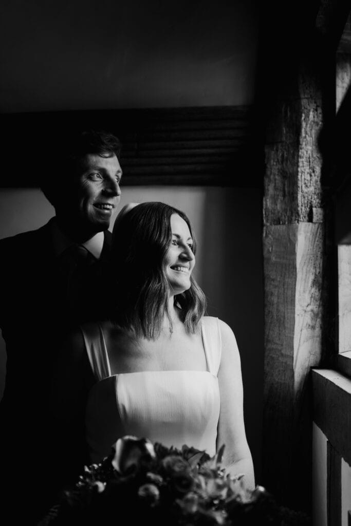 Romantic black and white wedding portrait at Birtsmorton Court. The bride and groom share a quiet moment by a historic timber-framed window, bathed in soft natural light. The bride, in a classic square-neck gown, smiles radiantly, holding her bouquet, while the groom looks on with joy. A stunning winter wedding in the Cotswolds, beautifully captured in timeless documentary wedding photography.