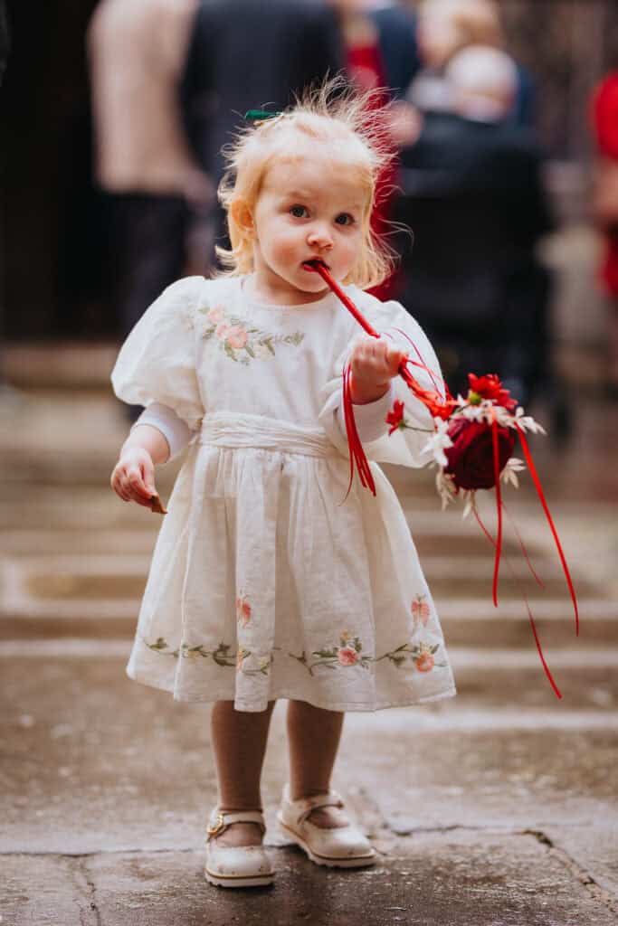 Adorable flower girl at a winter wedding at Birtsmorton Court. Dressed in a vintage-style embroidered dress with puff sleeves, she playfully chews on a red ribbon attached to her floral pomander. A beautifully candid moment capturing the innocence and charm of young wedding guests. A perfect addition to any Cotswolds wedding story, highlighting natural and documentary-style photography.