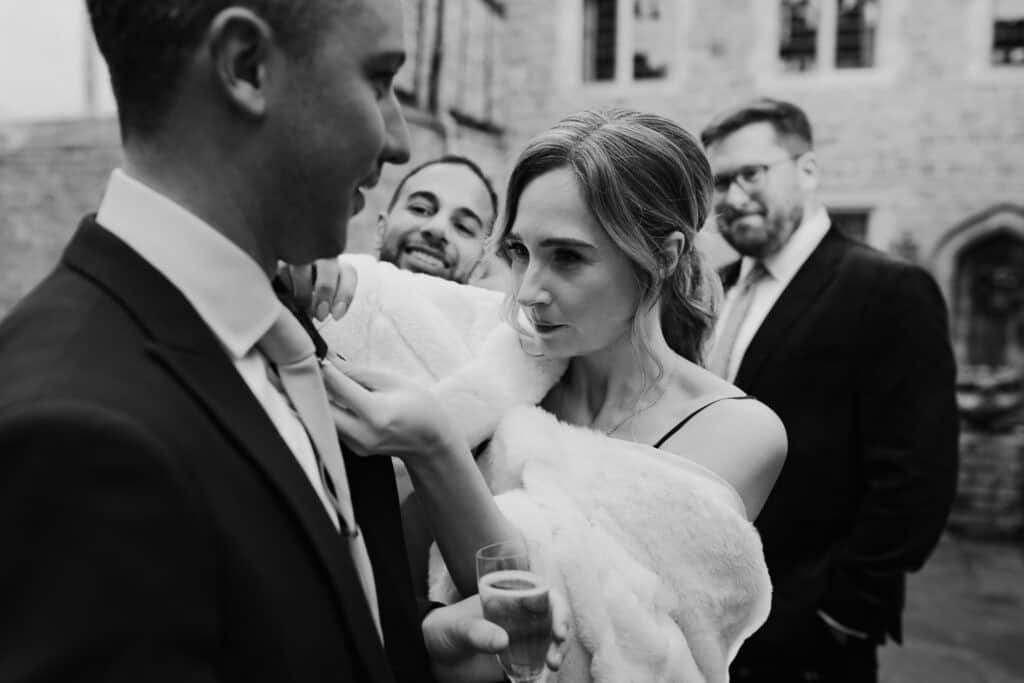 Candid wedding moment at Birtsmorton Court – a bridesmaid wrapped in a luxurious faux fur stole adjusts a groomsman’s tie with a focused expression, champagne flute in hand. Laughter and warmth fill the scene as wedding guests enjoy the winter celebration. A perfect example of natural and documentary-style wedding photography, capturing the connections and emotions of a Cotswolds wedding