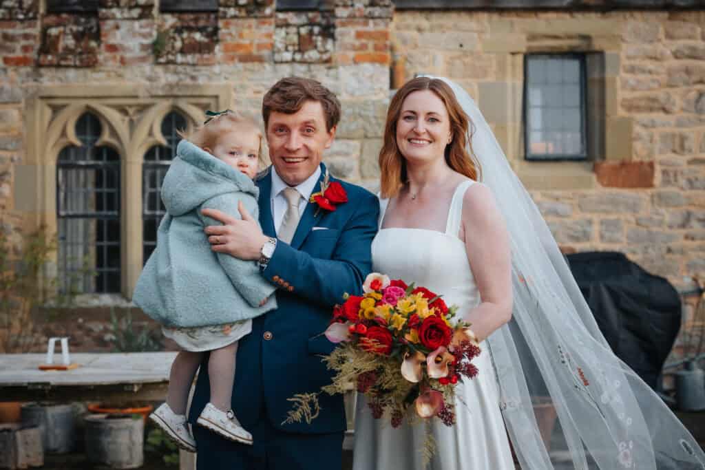 Beautiful family portrait at Birtsmorton Court – the newlyweds share a joyful moment with their young daughter against the stunning backdrop of the historic Cotswolds venue. The groom, dressed in a tailored blue suit, holds their little one wrapped in a cosy winter coat, while the bride beams in an elegant square-neck wedding gown, holding a vibrant bouquet of red and gold florals. A perfect example of relaxed and natural wedding photography capturing authentic emotions at this magical winter wedding.