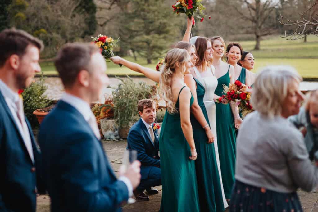 Playful wedding moment at Birtsmorton Court – the groom sneaks behind the bridesmaids, crouching down to hold his new wife's dress as they celebrate with bouquets raised high. The bride, glowing in her elegant white gown, stands among her bridesmaids dressed in rich emerald green, creating a stunning contrast against the picturesque Cotswolds backdrop. A fun and candid wedding photo capturing laughter, love, and unforgettable memories at this winter wedding celebration.