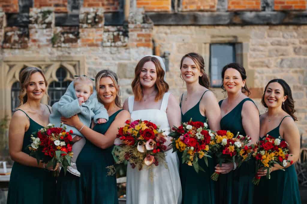 Elegant bridal party at Birtsmorton Court – the bride radiates joy surrounded by her bridesmaids dressed in stunning emerald green gowns, each holding vibrant red and gold winter bouquets. A heartwarming moment as one bridesmaid holds a bundled-up little guest, adding a touch of warmth to this winter wedding in the Cotswolds. A perfect blend of laughter, friendship, and beautiful wedding photography capturing the magic of the day.
