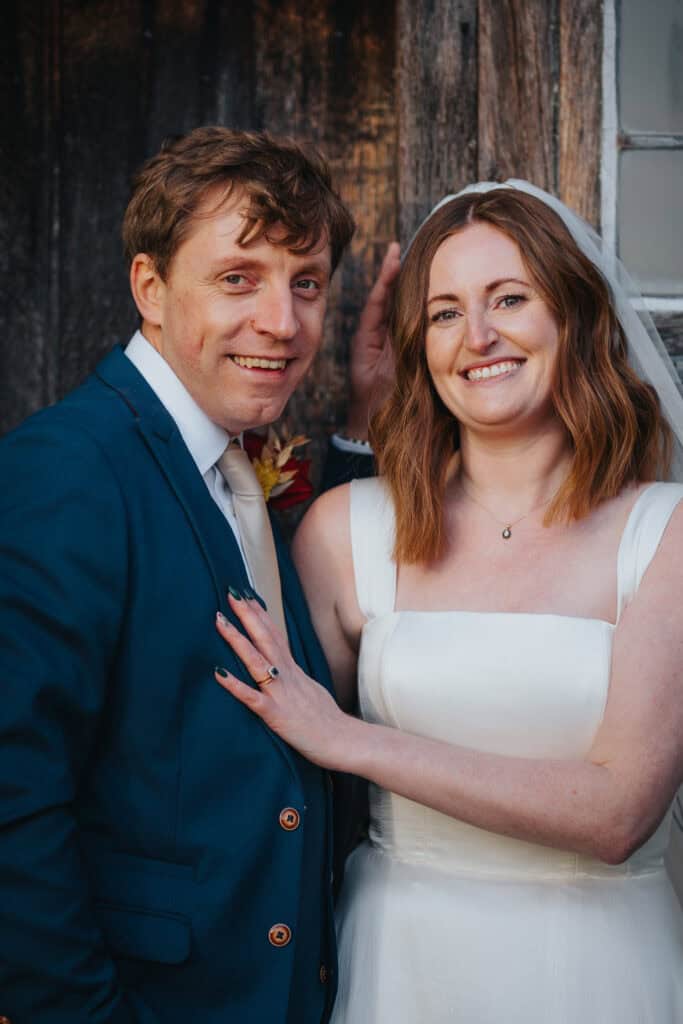 Joyful newlywed portrait at Birtsmorton Court – the bride and groom share a radiant smile against the rustic wooden backdrop of this stunning Cotswolds wedding venue. A timeless moment capturing love, elegance, and the beauty of a winter wedding celebration. Perfect inspiration for couples seeking natural and heartfelt wedding photography.