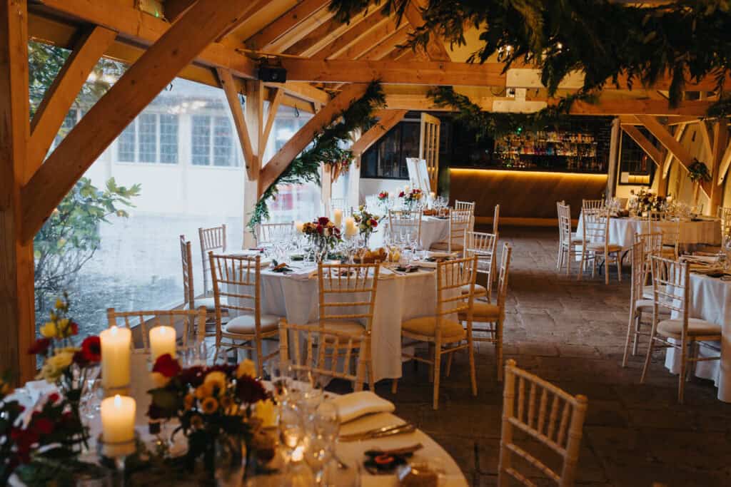 Elegant winter wedding reception at Birtsmorton Court – a beautifully decorated rustic barn setting with warm candlelight, floral centrepieces, and wooden beams adorned with greenery. A romantic and intimate atmosphere for a Cotswolds wedding celebration, perfect for couples seeking a cosy yet stylish venue.