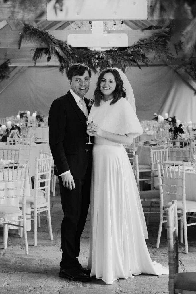 Elegant black and white wedding portrait of newlyweds at Birtsmorton Court. The couple stands in their beautifully decorated winter reception venue, surrounded by candlelit tables and festive greenery. A timeless Cotswolds wedding moment, perfect for couples looking for classic and emotive wedding photography inspiration.
