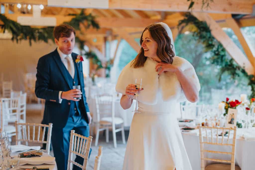 Candid moment between newlyweds at their winter wedding reception at Birtsmorton Court. The bride, wrapped in a luxurious white fur stole, shares a joyful glance while holding a glass of champagne, as the groom follows behind. A beautifully decorated Cotswolds wedding venue with festive florals and elegant table settings creates the perfect backdrop for this magical December wedding.