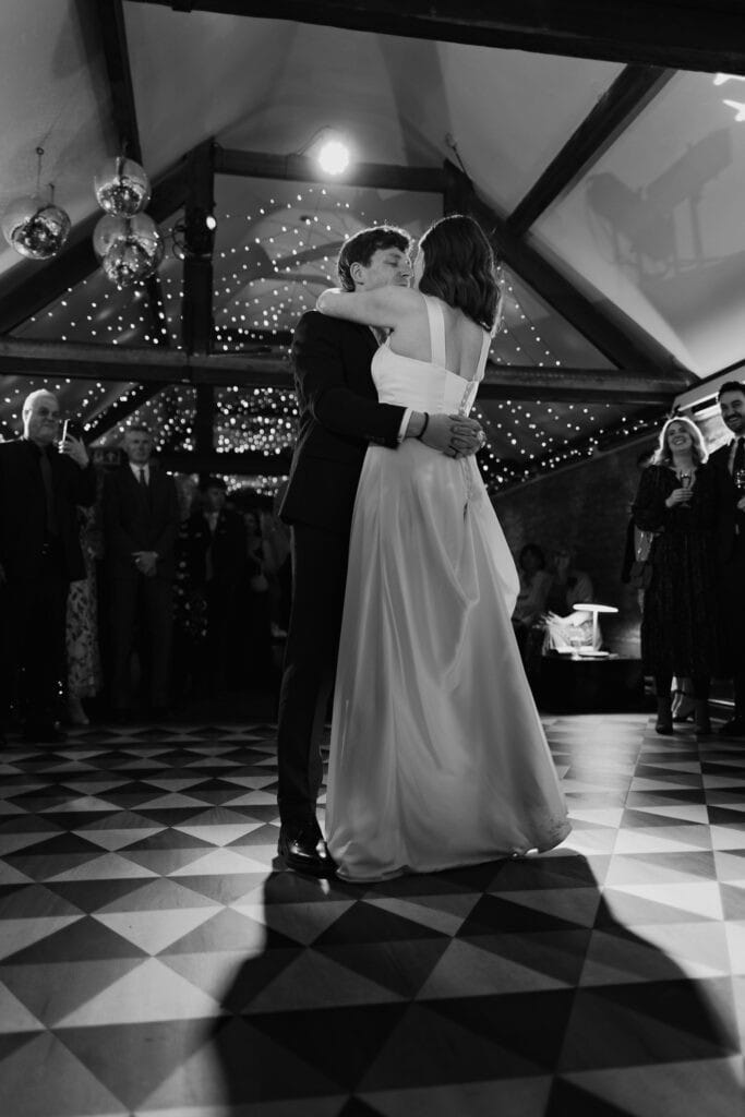 Elegant first dance at Birtsmorton Court – the newlyweds share an intimate embrace beneath twinkling fairy lights, creating a timeless and romantic moment. A beautifully captured black and white wedding photograph showcasing the magic of a Cotswolds winter wedding.