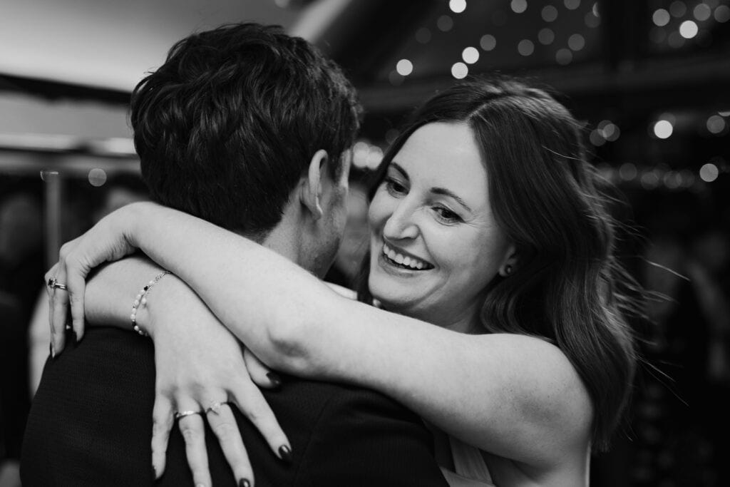 Emotional first dance at Birtsmorton Court – a stunning black and white wedding photograph capturing the bride's radiant smile as she embraces her groom. The intimate moment is set against twinkling fairy lights, making for a timeless and romantic winter wedding memory in the Cotswolds.
