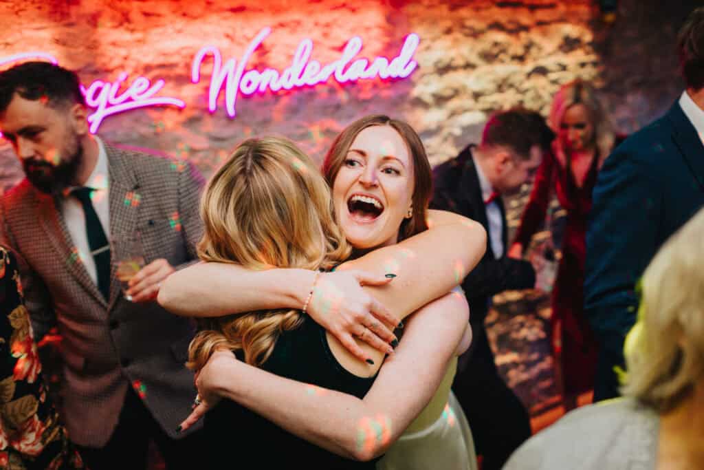 Joyful embrace on the dance floor at Birtsmorton Court – the bride shares a heartfelt moment with a guest during the evening wedding party. Neon lights, music, and laughter fill the air, capturing the energy of a fun-filled winter wedding celebration in the Cotswolds.