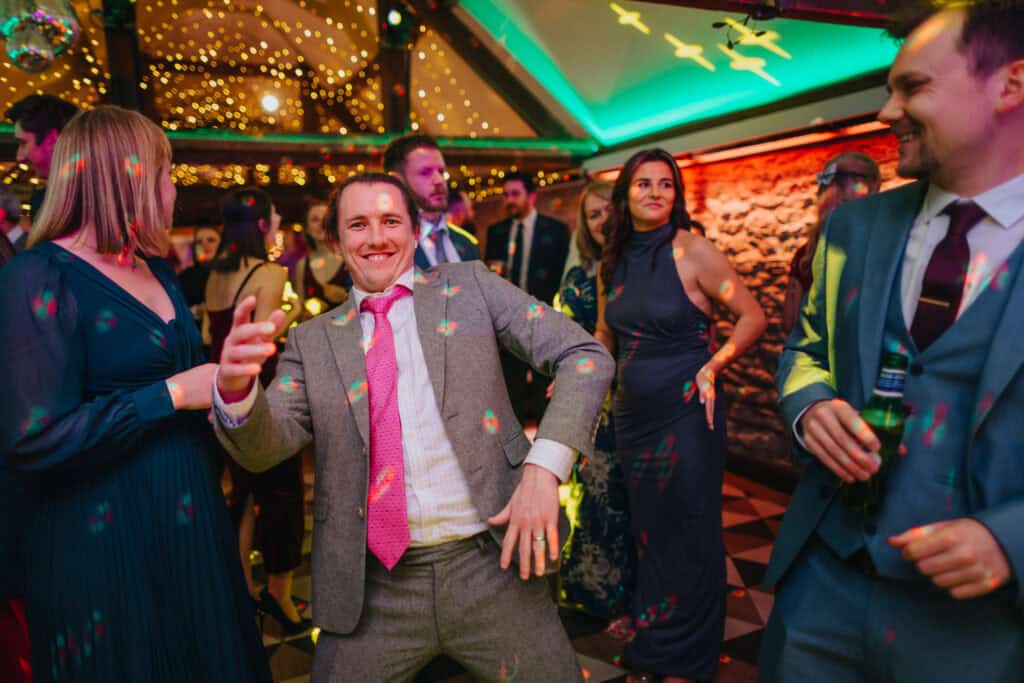 Fun and lively wedding reception at Birtsmorton Court with guests dancing under colourful lights. A guest in a grey suit and pink tie steals the show on the dance floor, capturing the energy and joy of the evening party at this stunning winter wedding in the Cotswolds.