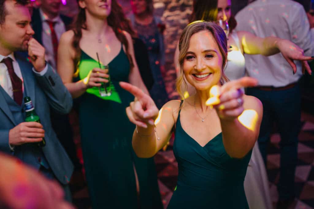 Vibrant evening party at Birtsmorton Court with guests celebrating on the dance floor. A bridesmaid in an elegant green dress beams with joy, dancing under colourful lights at this magical winter wedding in the Cotswolds.