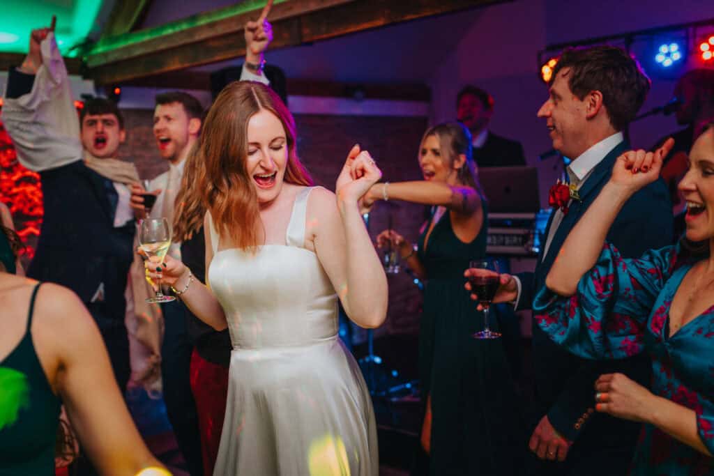 Joyful bride dancing at her evening wedding reception at Birtsmorton Court. Surrounded by friends and family, she celebrates with a glass of wine under vibrant party lights, making for an unforgettable Cotswolds winter wedding moment.