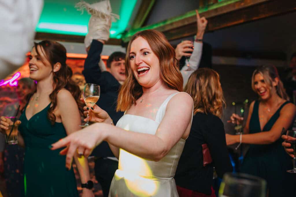 Radiant bride dancing and celebrating with friends at her evening wedding reception at Birtsmorton Court. Capturing the energy and joy of a winter wedding in the Cotswolds, surrounded by guests enjoying the party atmosphere with drinks and music.