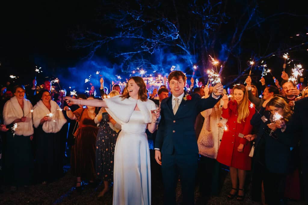 A magical winter wedding at Birtsmorton Court, with the newlyweds celebrating under a canopy of sparklers held by their guests. The bride, in a timeless white gown with a faux fur wrap, and the groom, in a classic navy suit, beam with joy as they lead the sparkler send-off. The warm glow of the sparklers contrasts beautifully with the deep blue night sky, creating an enchanting atmosphere. A perfect moment of celebration, love, and winter romance.