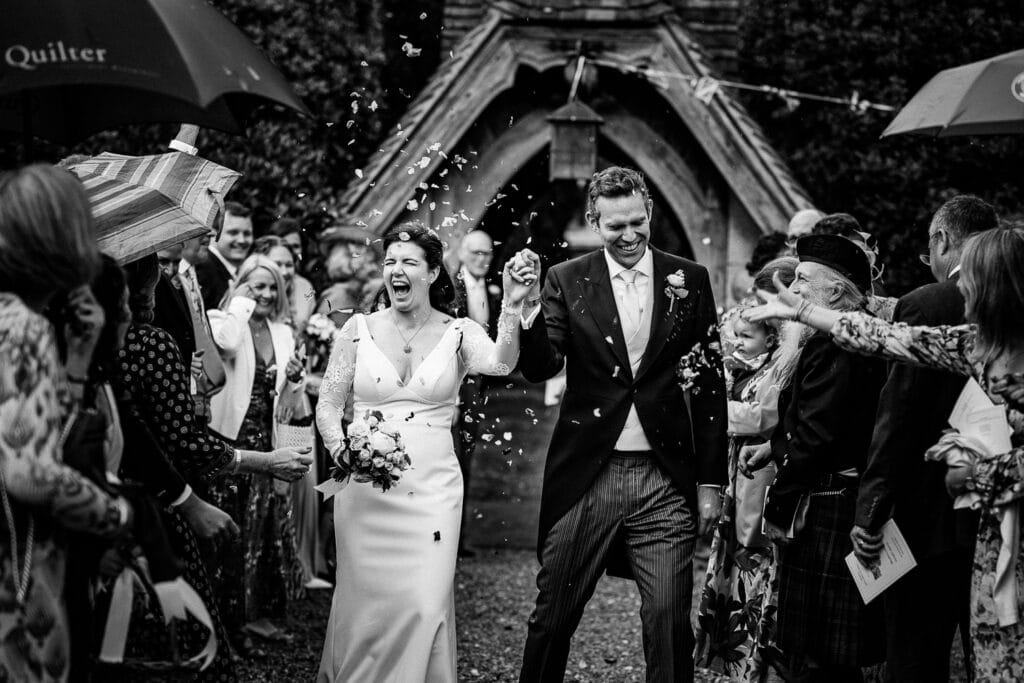 Black and white wedding photograph of a joyful bride and groom walking hand in hand through a confetti toss outside a historic church. The bride, wearing a fitted gown with lace sleeves, laughs with excitement while holding a bouquet, while the groom, dressed in a formal suit with a boutonnière, smiles warmly. Guests line both sides, throwing petals and celebrating the newlyweds in a candid, emotive moment.