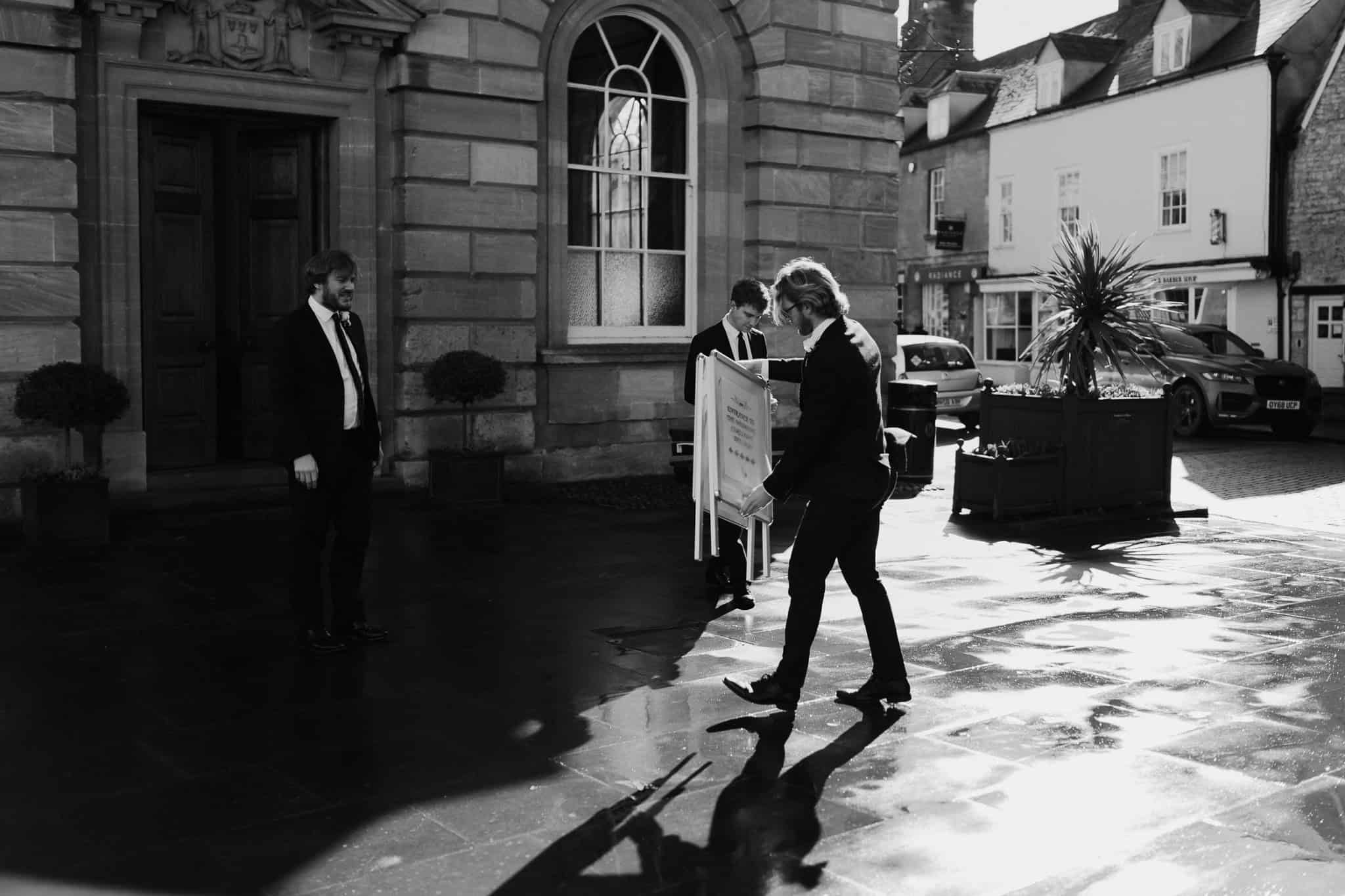 Black and white candid shot of groomsmen setting up outside Woodstock Town Hall before the wedding ceremony. A documentary-style wedding photo capturing authentic pre-ceremony moments.